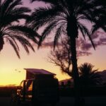 camper van at purple and yellow sunset parked between palm trees