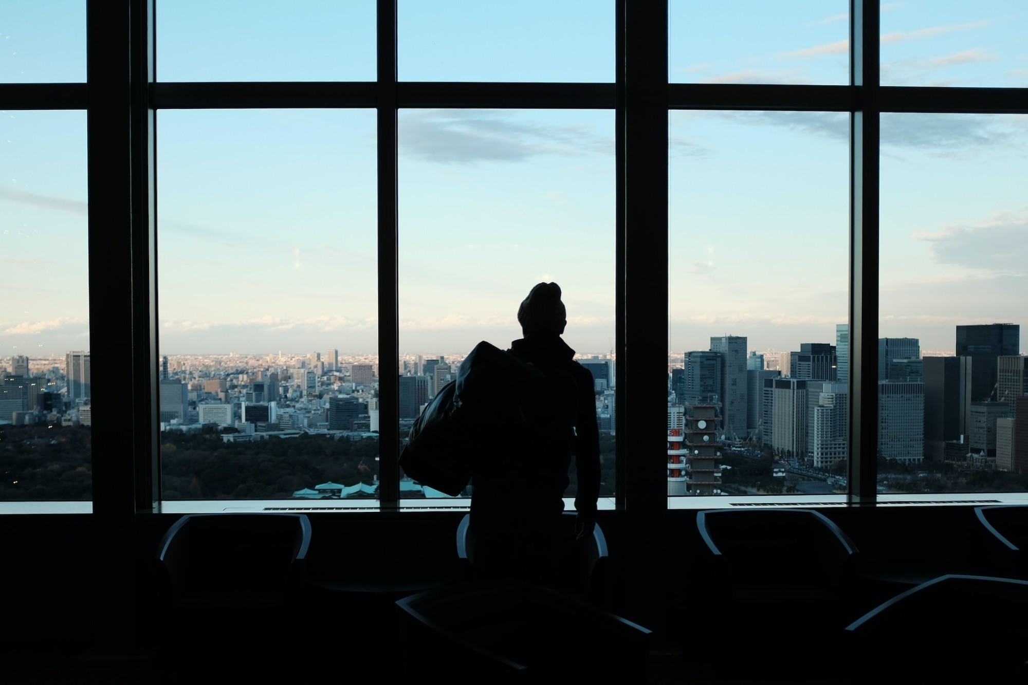 A man gazes out of an airport window at a city skyline, contemplating the global nature of corporate travel.