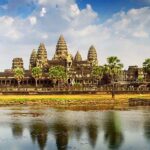 Angkor Wat at sunrise, reflecting in the water, with lush green surroundings, showcasing the ancient temple's grandeur and serene atmosphere.