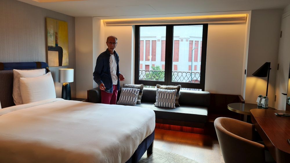 hotel room interior: bed, sofa, desk, table, window behind, and a man standing between the bed and the sofa.