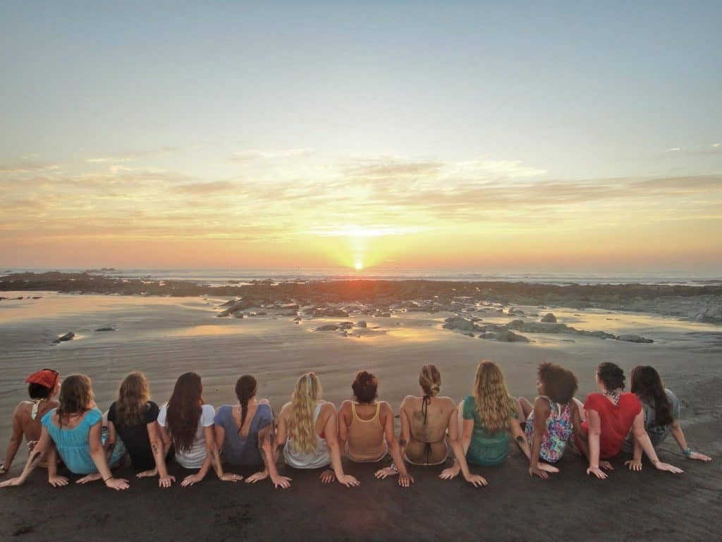 Twelve young women sitting next to each other on a beach looking toward the ocean at sunset.