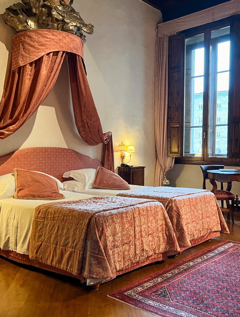 A luxurious bedroom featuring a traditional bed with a coronet and drapes, positioned by a large window with wooden shutters.