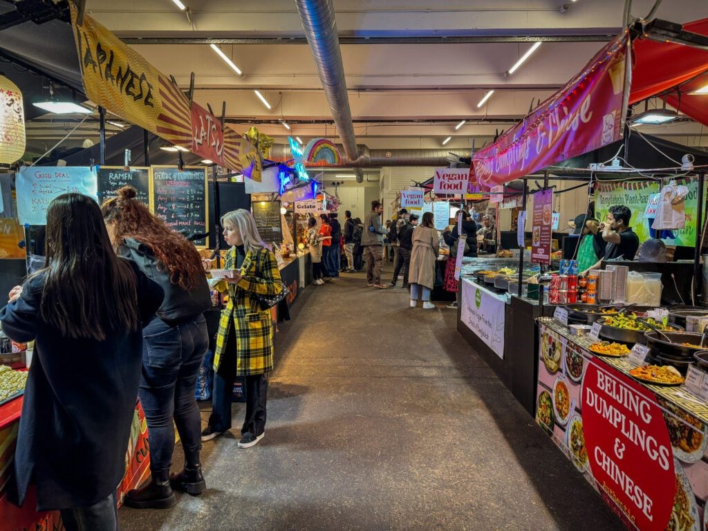 A vibrant street food market scene in Brick Lane, London, with diverse food stalls and bustling crowds