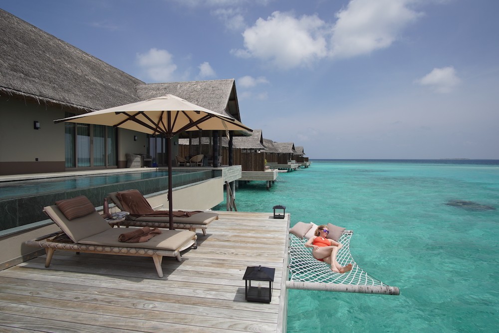 Luxurious overwater bungalows at the Joali resort in the Maldives, featuring private decks with cantilevered hammocks extending over the crystal-clear turquoise water, under a sunny sky, exemplifying a dreamlike tropical island escape