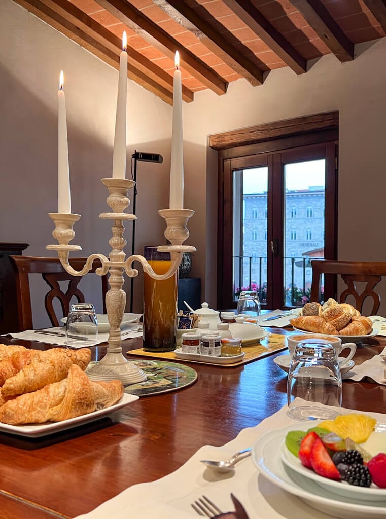 A formal dining table is elegantly set for breakfast with pastries, fresh fruit, and a silver candelabra casting a warm light.