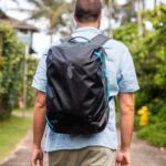 A person standing outside in a light blue short sleeve shirt wears the Cotopaxi Allpa 35L backpack, in black with a gray llama-head logo and aqua accents.