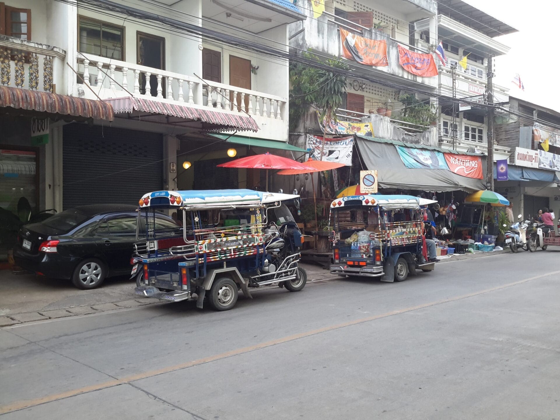 Street scene in Chiang Khong, Thailand, featuring two tuk-tuks parked in front of a row of local shops.