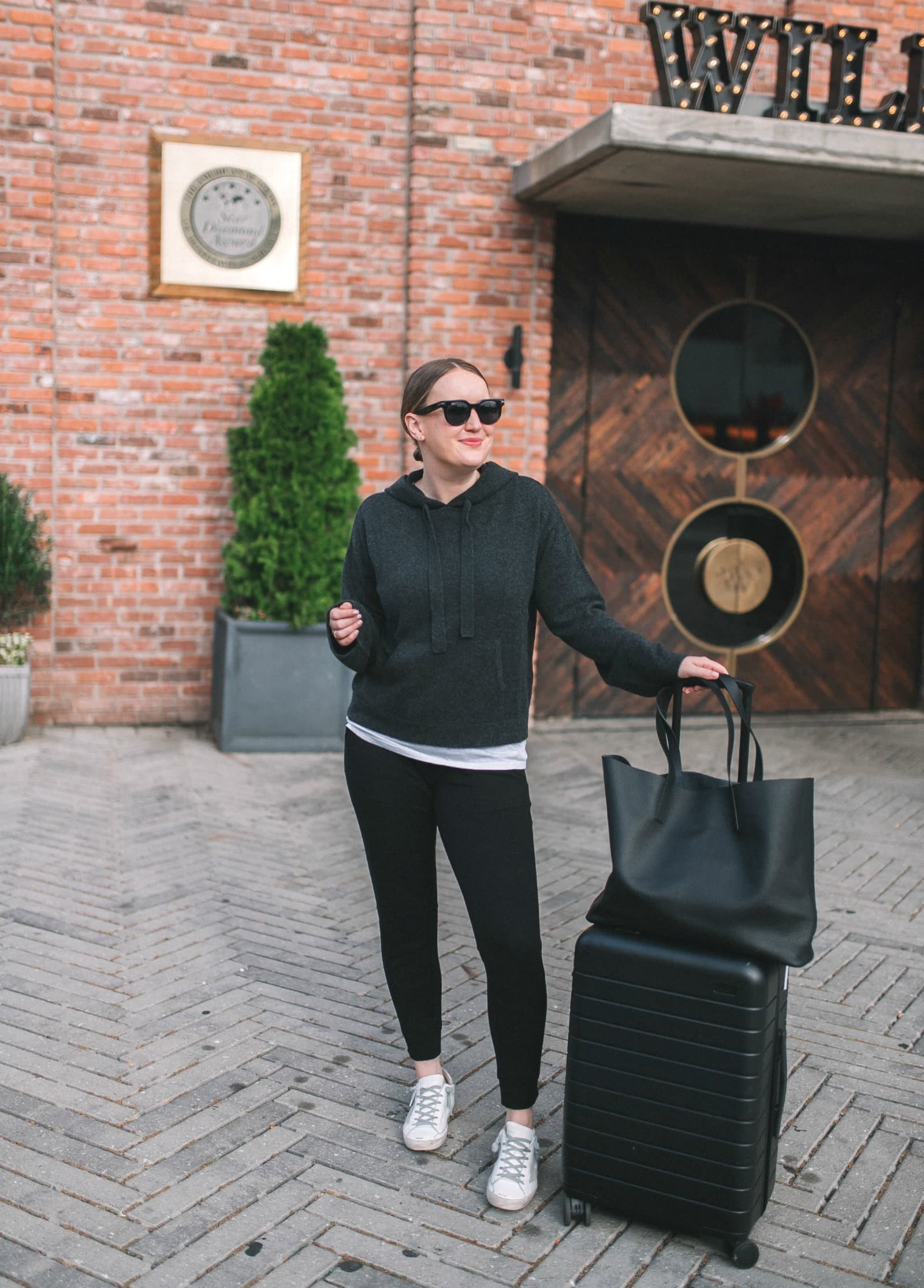 Woman in a cozy grey sweater, black joggers, and white sneakers, sitting in an airport lounge, smiling, representing comfortable yet stylish travel attire for long flights