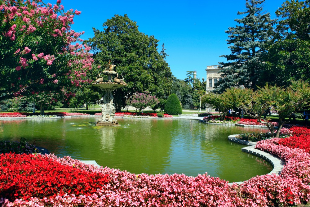 Gardens and flowers surrounding a tranquil pond at Dolmabahce Palace in Istanbul, Turkey, showcasing vibrant spring blooms, lush greenery, and Ottoman architecture, reflecting the city's blend of history and natural beauty