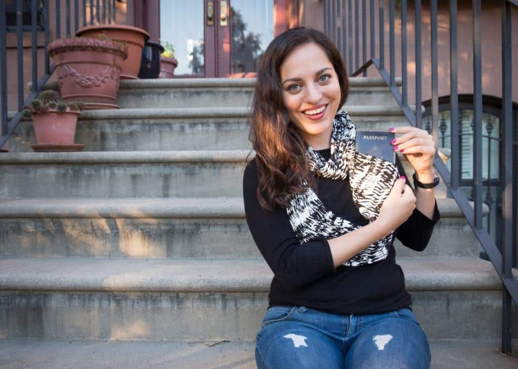 Kate sitting on a stoop in Brooklyn, wearing a circle scarf and showing that her passport is in the pocket.