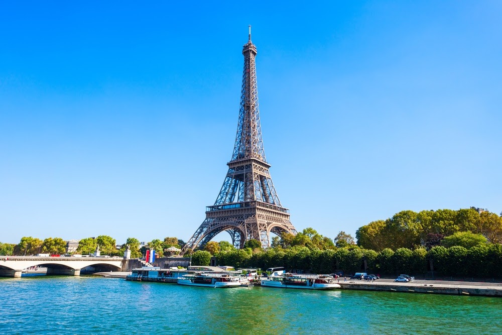 The iconic Eiffel Tower in Paris, France, majestically rising above the city skyline, with the River Seine flowing gently in the foreground, and blossoming trees adding a touch of spring color to the urban landscape