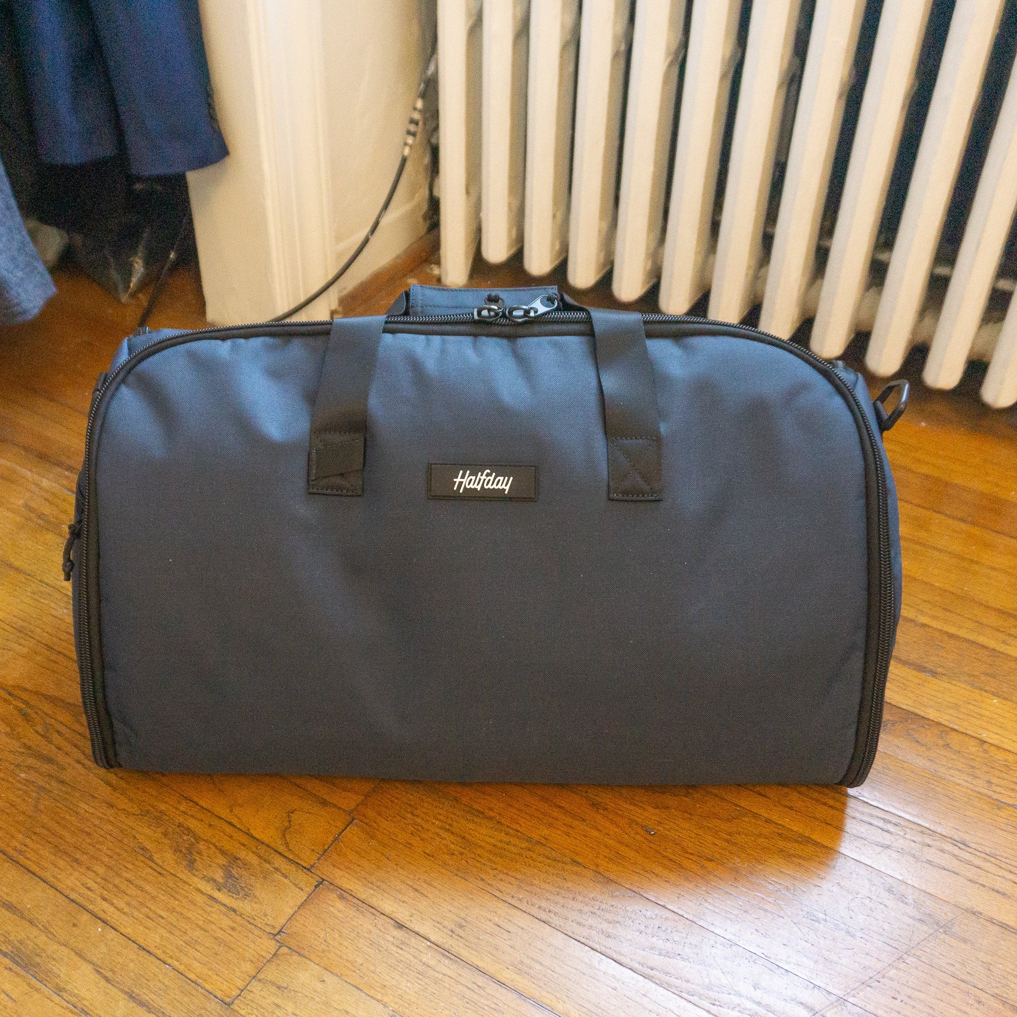 Halfday Garment Duffel bag in a neutral color, placed on a light wooden floor, showcasing its exterior design.