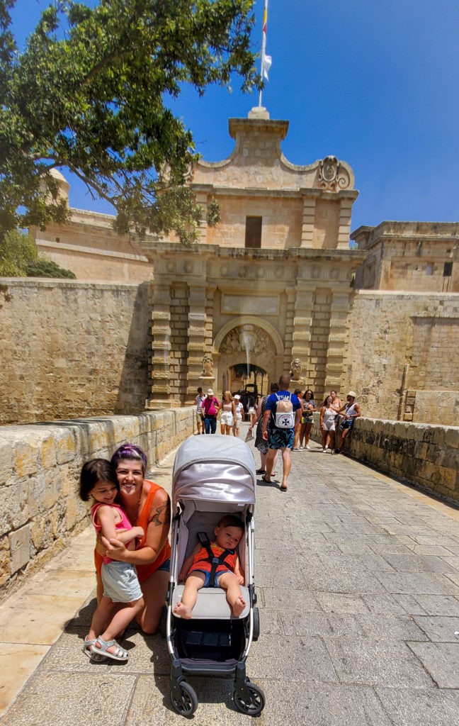 woman hugging toddler and baby in stroller in front of gate of mdina