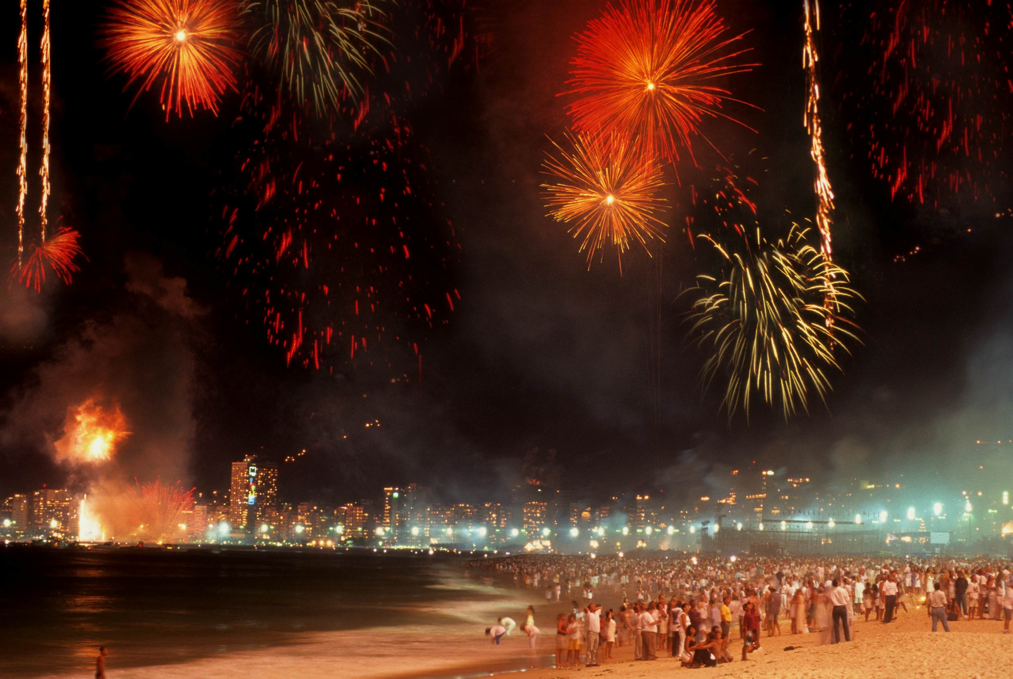 New Year's Eve fireworks illuminate Copacabana beach in Rio de Janeiro, with crowds gathered along the shore and buildings visible in the background.