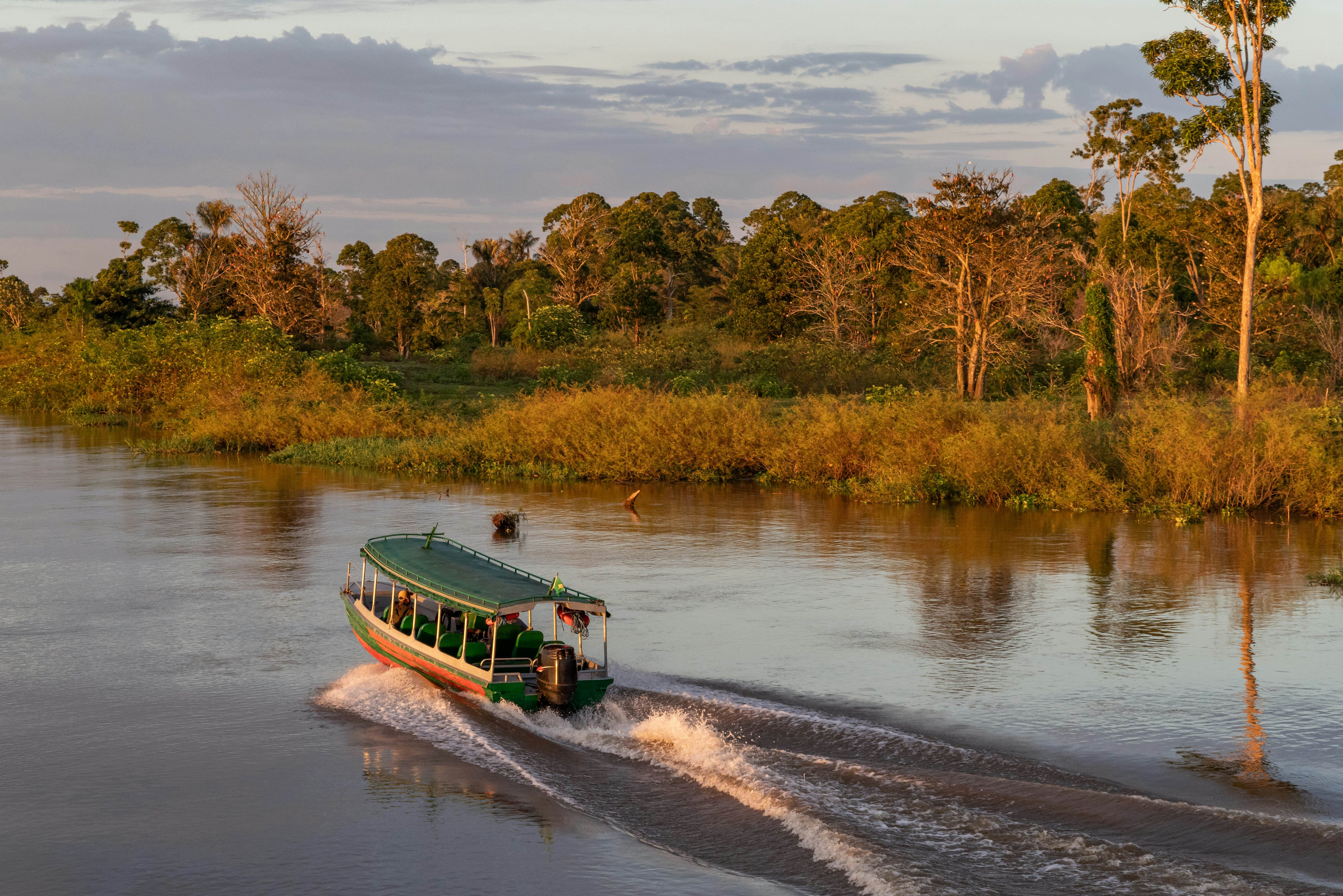 The Amazon River at sunrise, showing a boat leaving a wake and lush green rainforest lining the riverbanks under a golden sky.