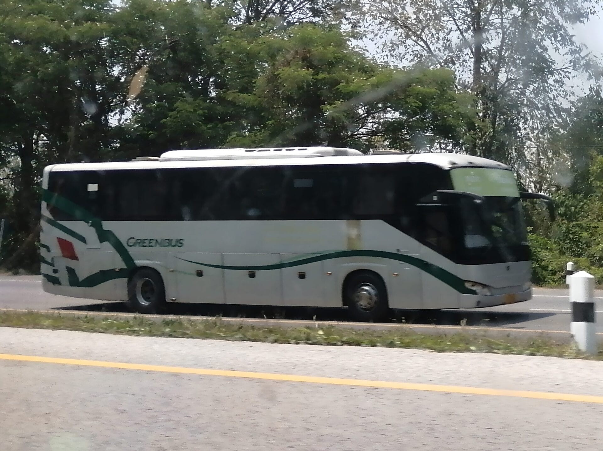 Greenbus driving on a road in Thailand with trees in the background.