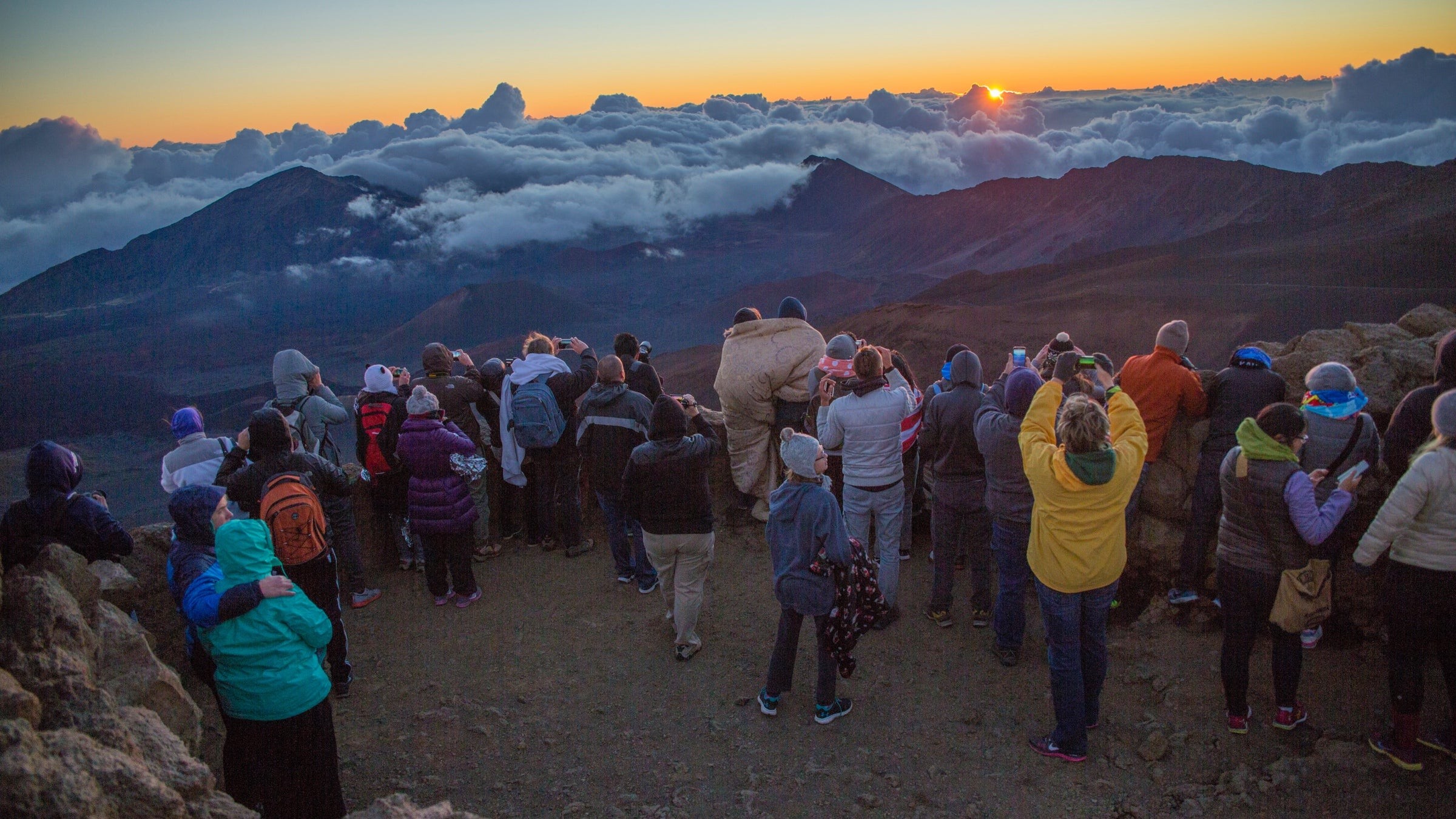 A crowd of people wearing jackets, some bundled in sleeping bags, watch the sunrise from atop Maui