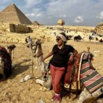 Woman in a blue long-sleeve shirt and beige capri pants standing in front of Egyptian pyramids, showcasing modest and practical travel wear for women.