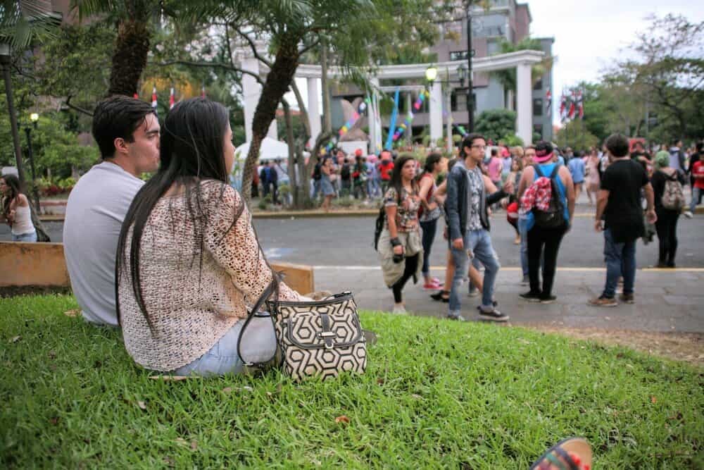 A couple on the foreground sitting on the grass in a city park, with more people walking on the sidewalk and street. 