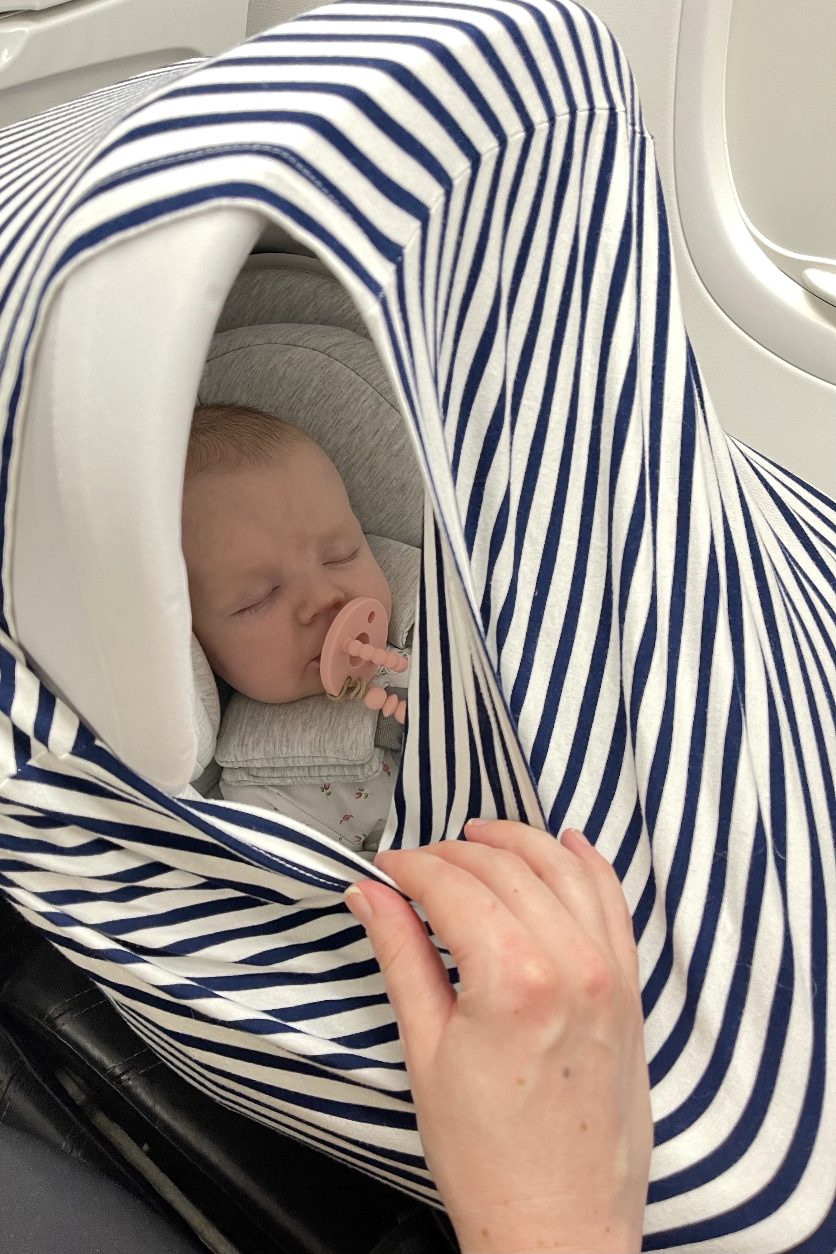 Baby napping in a car seat with a cover, demonstrating how to create a darker, quieter space for sleep on an airplane.