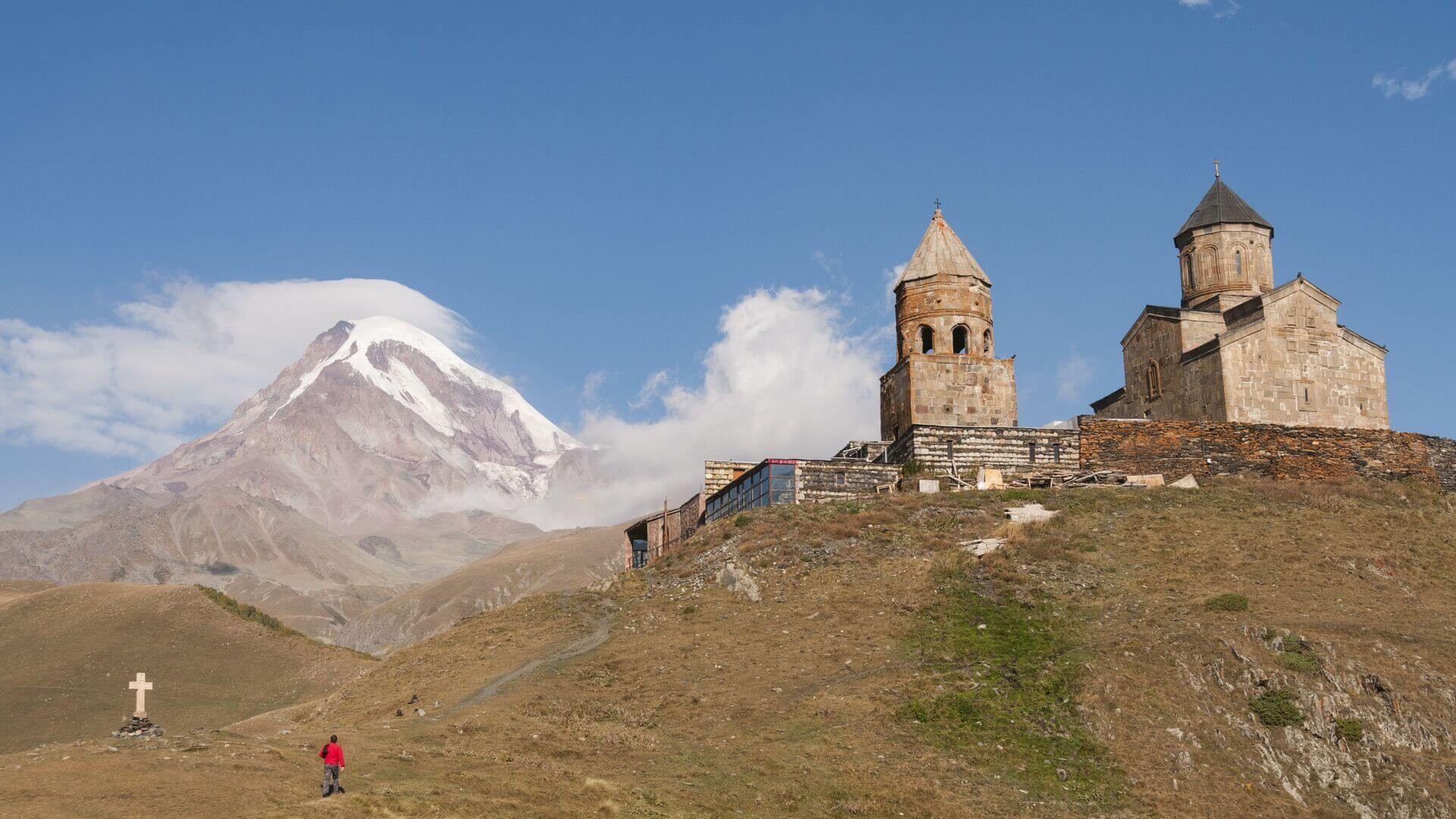 A solitary figure walking along a mountain road towards the iconic silhouette of Gergeti Trinity Church against the backdrop of the towering Caucasus Mountains.