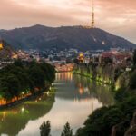 Panoramic view of Tbilisi at sunset with city lights reflecting on the Kura River, highlighting the blend of modern and historic architecture.