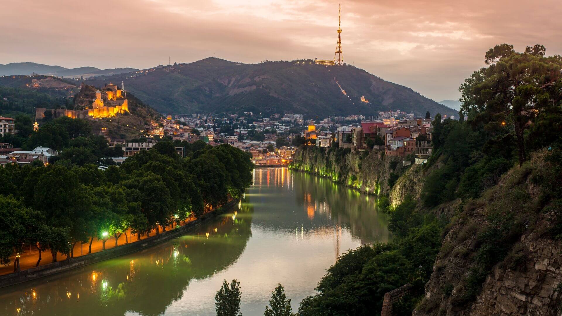 Panoramic view of Tbilisi at sunset with city lights reflecting on the Kura River, highlighting the blend of modern and historic architecture.