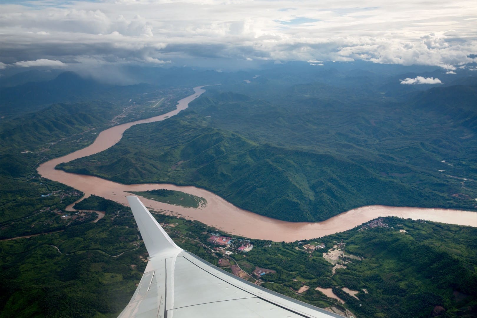 Aerial view of the landscape in Laos with a river winding through green mountains and an airplane wing visible in the foreground under a cloudy sky.