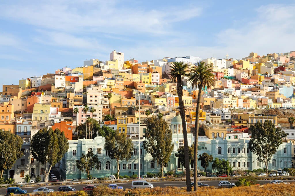 A vibrant hillside filled with colorful houses in Las Palmas, Gran Canaria, with two palm trees in the foreground under a sunny sky.