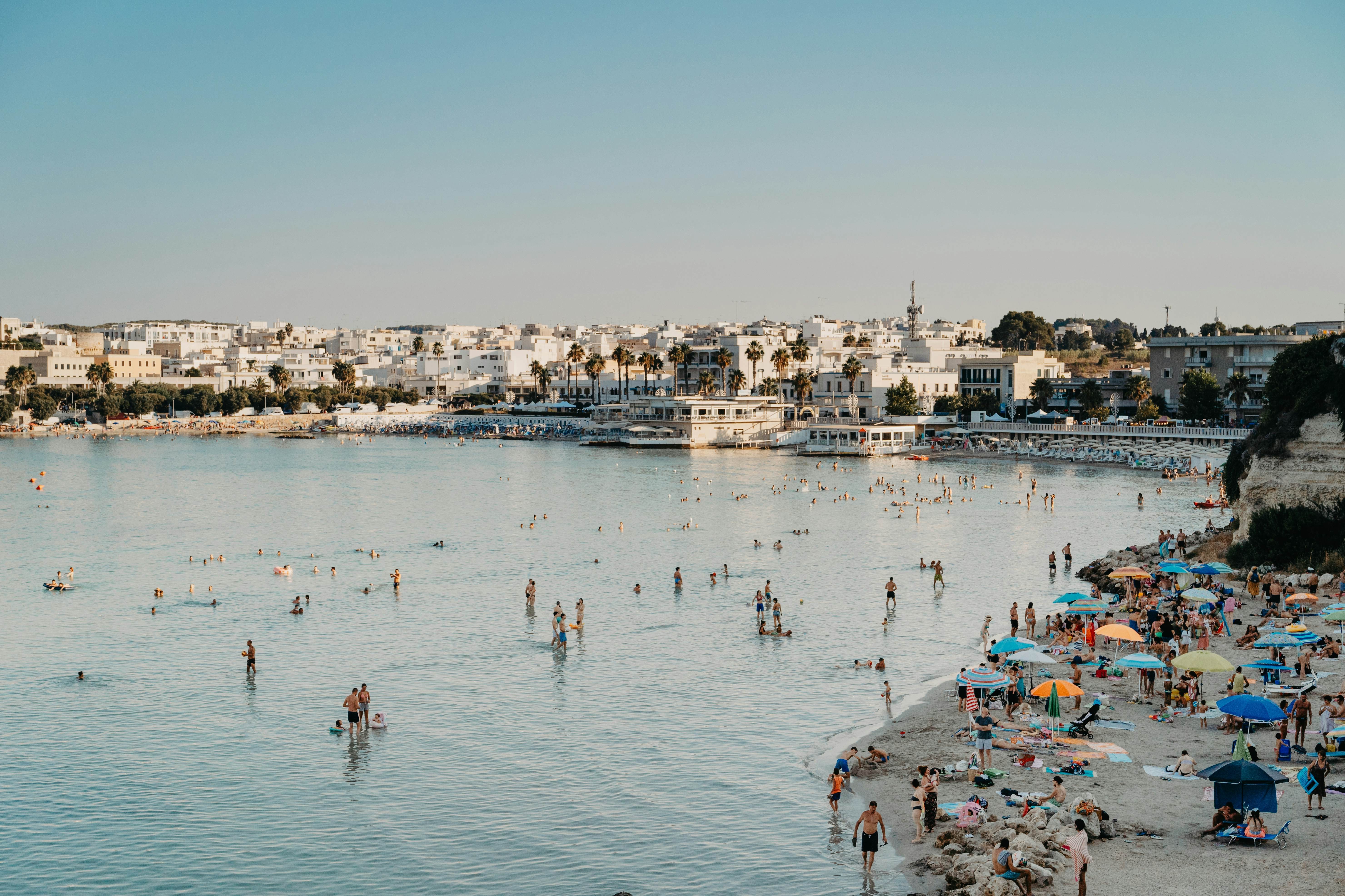 People crowd on a beach and swim in the sea with a whitewashed city in the background