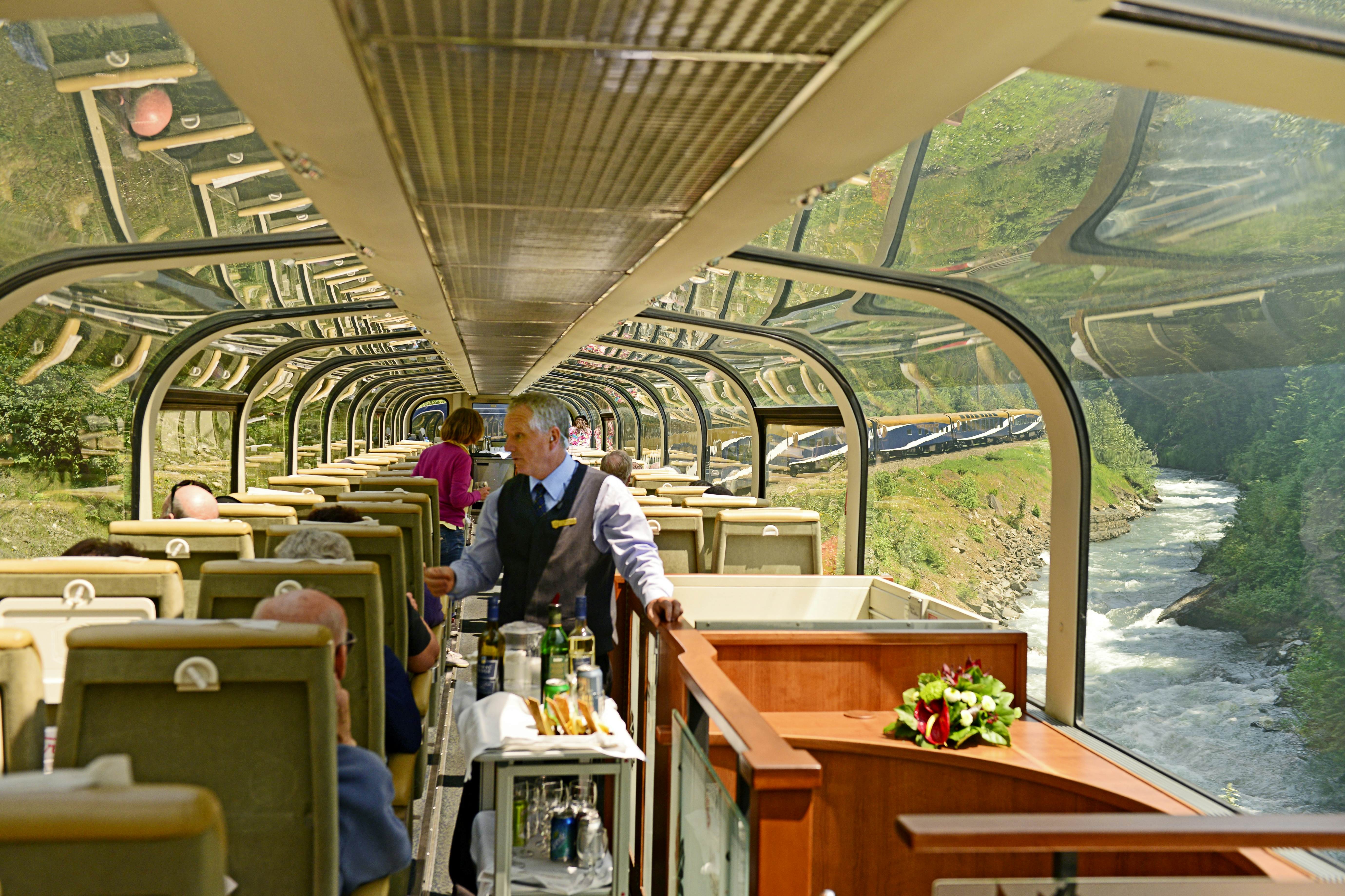 A train attendant wheels a drink cart down the aisle of an observation car. Passengers are seated enjoying views of a river and green mountains.