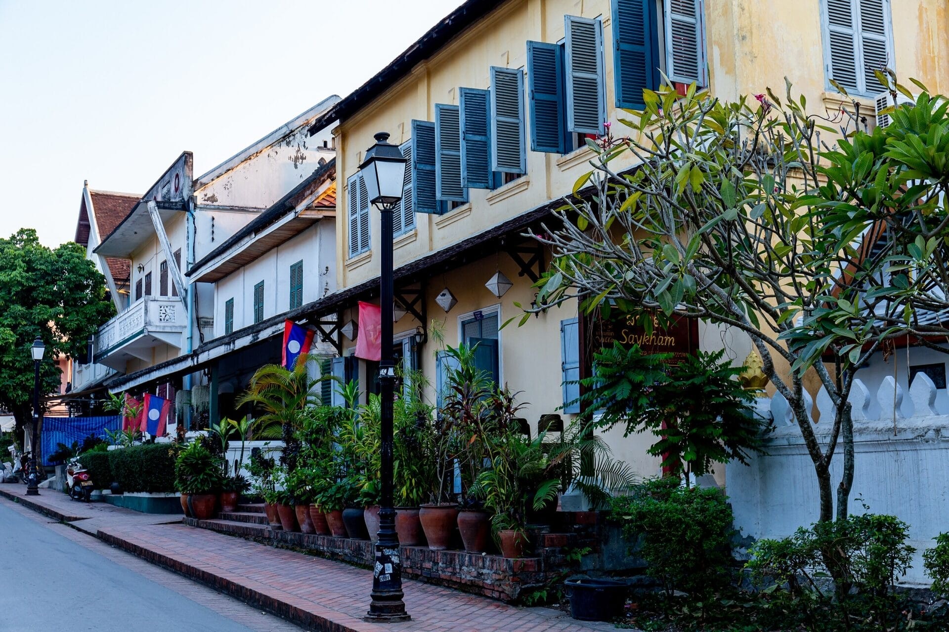 A picturesque street in Luang Prabang, Laos, lined with colonial-style buildings adorned with green shutters, potted plants, and national flags.