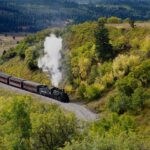 alt text: Rocky Mountaineer train with glass dome car traveling through red rock landscape