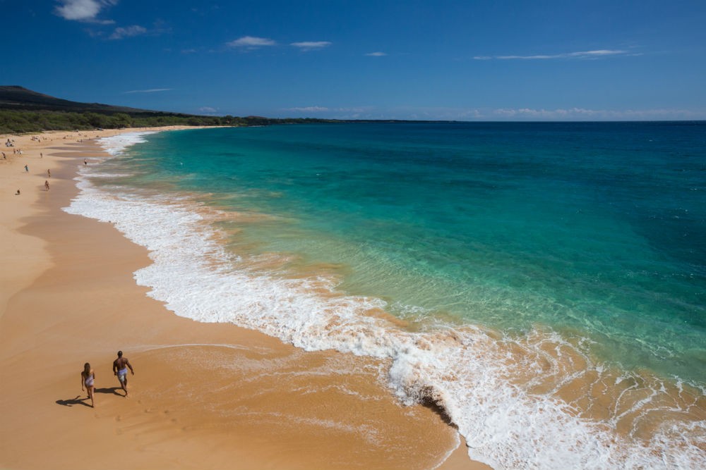 Makena beach in Maui, Hawaii, showcasing golden sand and clear blue water, with gentle waves lapping the shore, palm trees swaying in the background, and a clear sky above, perfect for a spring vacation