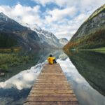 Man sitting on boat pier in front of scenic lake reflecting peaceful travel amidst global uncertainties