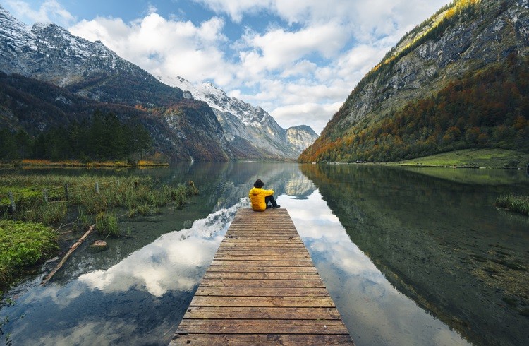 Man sitting on boat pier in front of scenic lake reflecting peaceful travel amidst global uncertainties