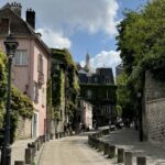 A winding street in Montmartre, Paris, featuring a stone wall on the right and the iconic pink house covered in ivy on the left.