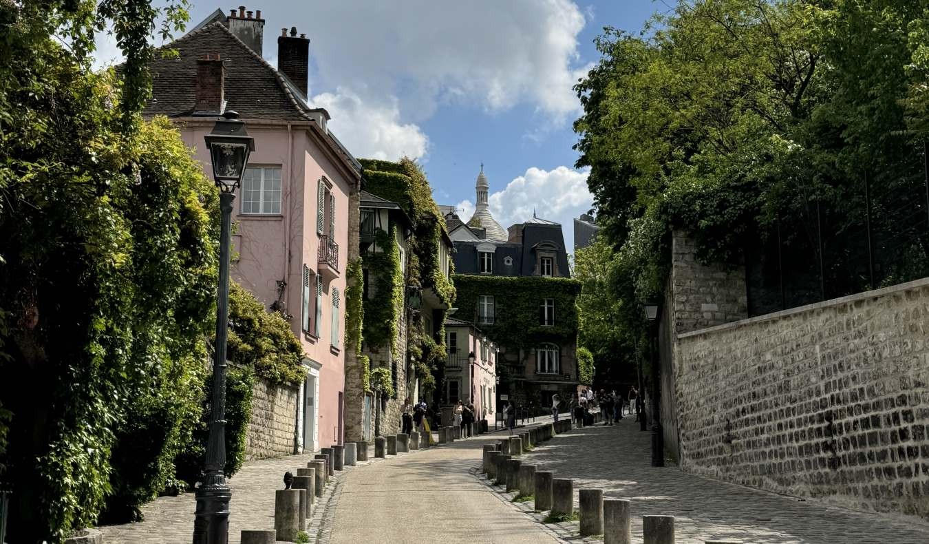 A winding street in Montmartre, Paris, featuring a stone wall on the right and the iconic pink house covered in ivy on the left.