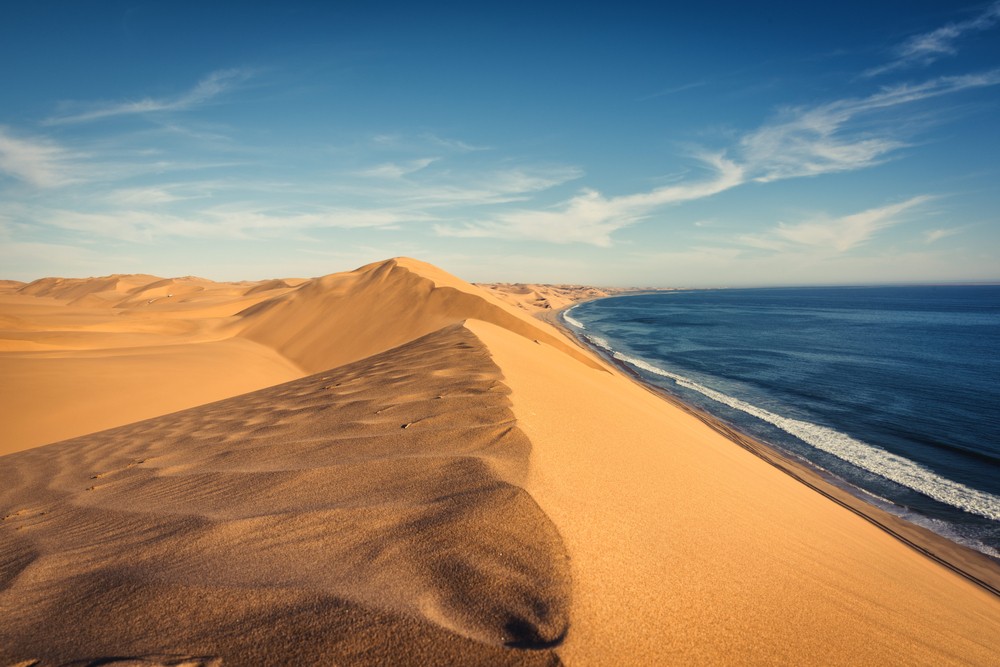 The vast and dramatic Namib Desert in Namibia, stretching to the Atlantic Ocean coastline, featuring towering sand dunes meeting the ocean waves, under a clear blue sky, showcasing the unique and breathtaking desert landscape