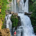 A woman wearing a red two-piece swim suit in posing on a boulder in front of a tall two-tiered waterfall