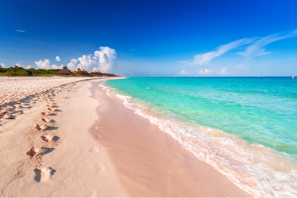 A serene and empty beach in Playa del Carmen, Riviera Maya, Mexico, featuring soft white sand, turquoise Caribbean waters, and gentle footprints leading towards the horizon under a bright sky, illustrating the tranquility of a spring getaway