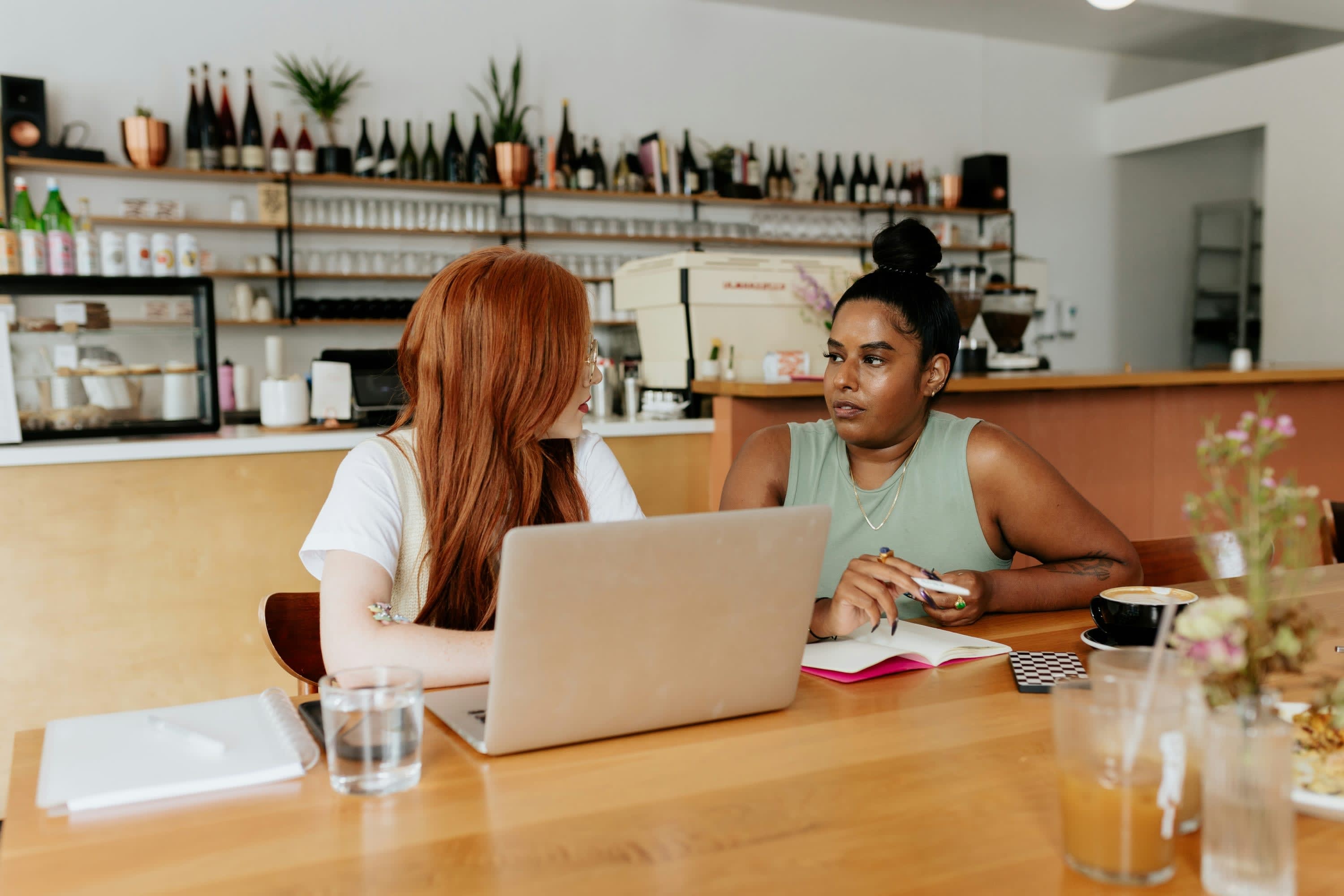 Two women collaborate on some type of work while drinking coffee at a coffee bar