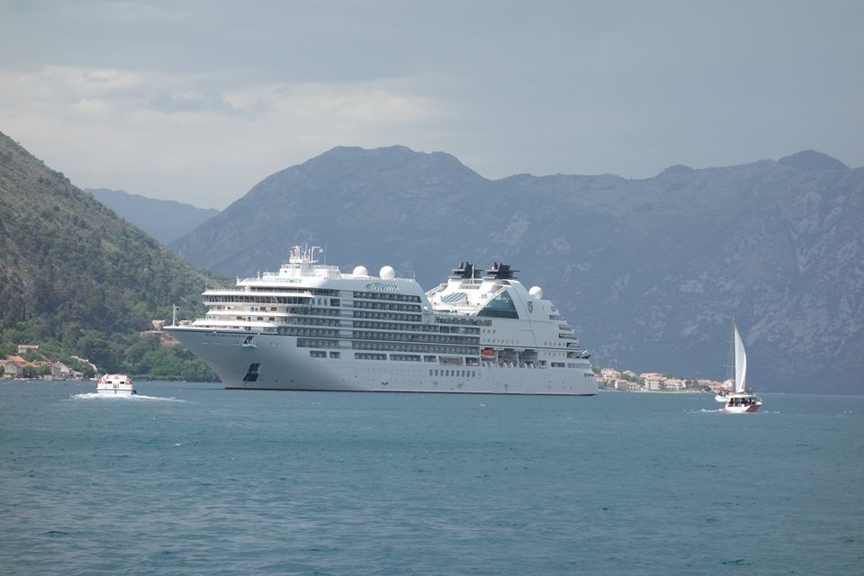 The elegant Seabourn Ovation cruise ship sailing on calm blue waters, with picturesque coastal mountains in the background, under a clear sky, representing the luxury and scenic beauty of a Mediterranean cruise in spring