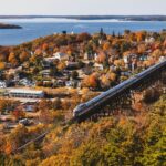 An aerial view captures a train gracefully traversing a long trestle bridge, with a picturesque waterfront town nestled amidst lush trees in vibrant fall foliage.