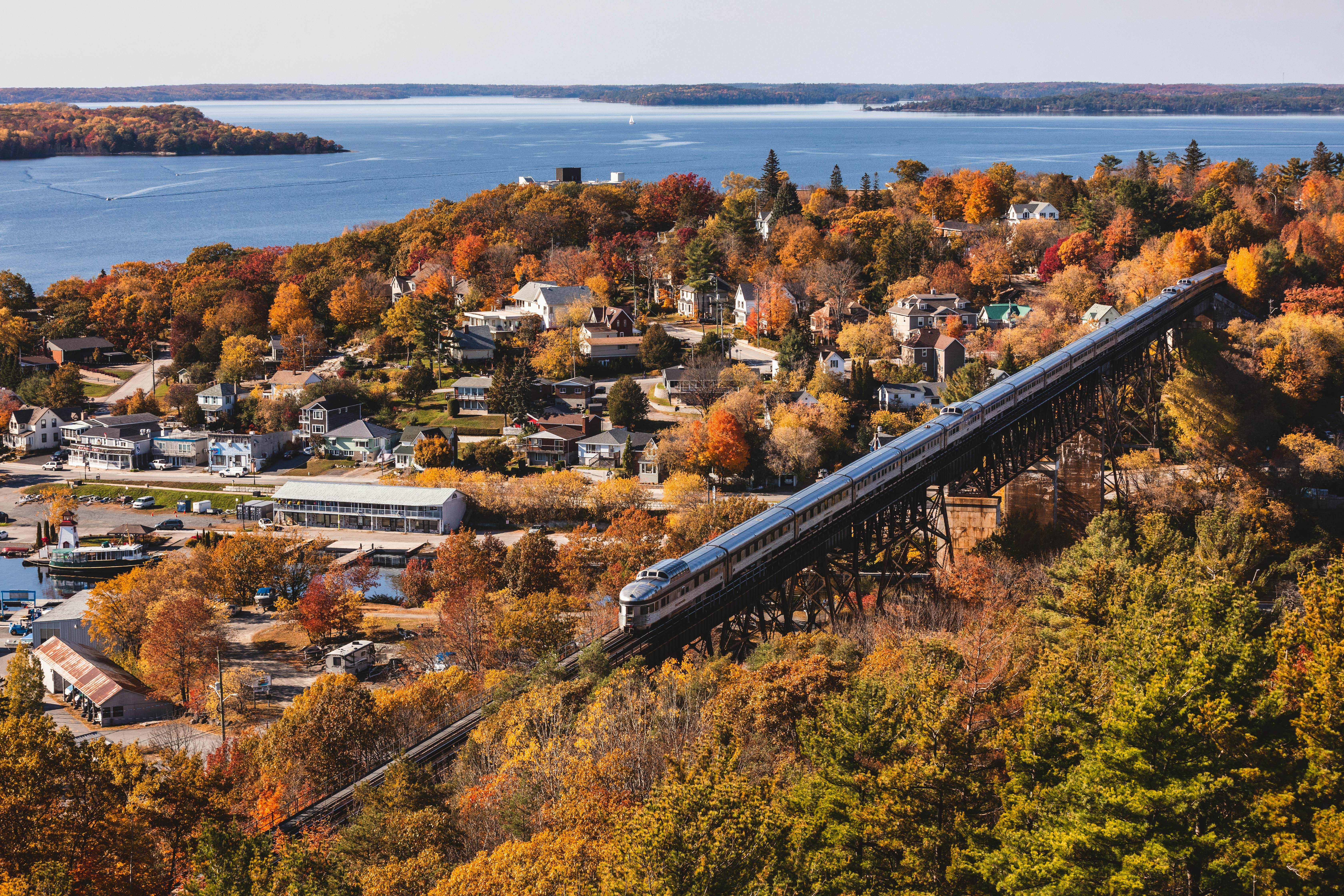 An aerial view captures a train gracefully traversing a long trestle bridge, with a picturesque waterfront town nestled amidst lush trees in vibrant fall foliage.