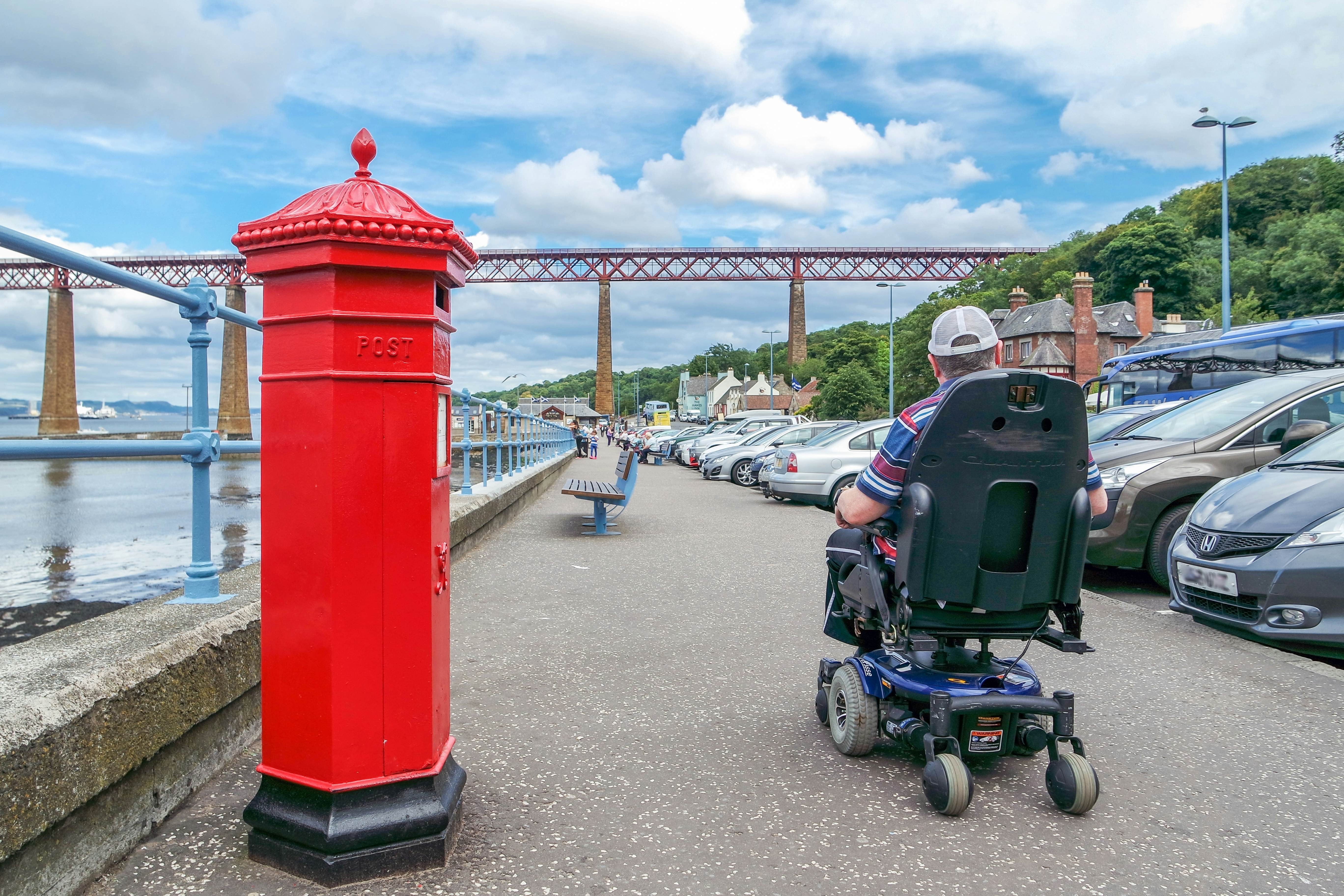 A man in wheelchair passes a letter box by the Firth of Forth, near the Forth Bridge, Edinburgh, Scotland