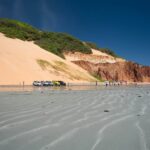 Beachgoers relax on the sandy shore of Porto de Galinhas, Pernambuco, with calm, shallow turquoise water and a jangada sailboat visible.