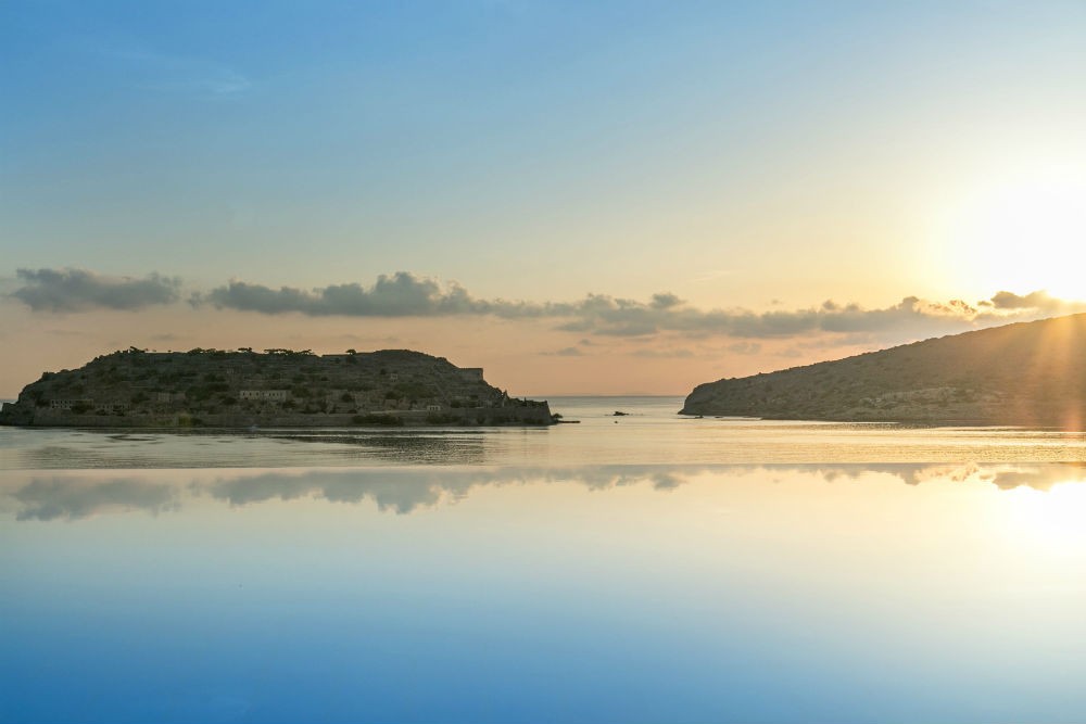 Spinalonga Island, Crete, Greece, a historic fortress island just off the coast, with clear turquoise waters surrounding it and the mainland of Crete visible in the distance, under a sunny sky, showcasing a blend of history and natural beauty