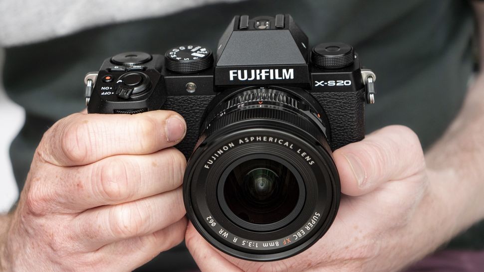 A hand holding the Fujifilm X-S20 camera, showcasing its grip and screen in an outdoor setting, highlighting its suitability for travel photography