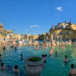 View of the open air thermal baths in Budapest with many people enjoying the warm water under a clear blue sky.